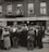 A large crowd of people stands gathered outside a brick building with signs for veteran bonus applications and music services. Many individuals wear hats and coats typical of the early 20th century. Some people can be seen on the balconies and looking out from windows, observing the crowd below.