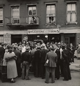 A large crowd of people stands gathered outside a brick building with signs for veteran bonus applications and music services. Many individuals wear hats and coats typical of the early 20th century. Some people can be seen on the balconies and looking out from windows, observing the crowd below.