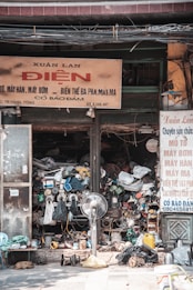 A cluttered and busy storefront filled with various mechanical and electrical items stacked chaotically. A large fan is placed in the center, surrounded by numerous wires, tools, and parts. Signs in a foreign language are displayed above and beside the shop entrance, indicating services related to repairs and machinery.