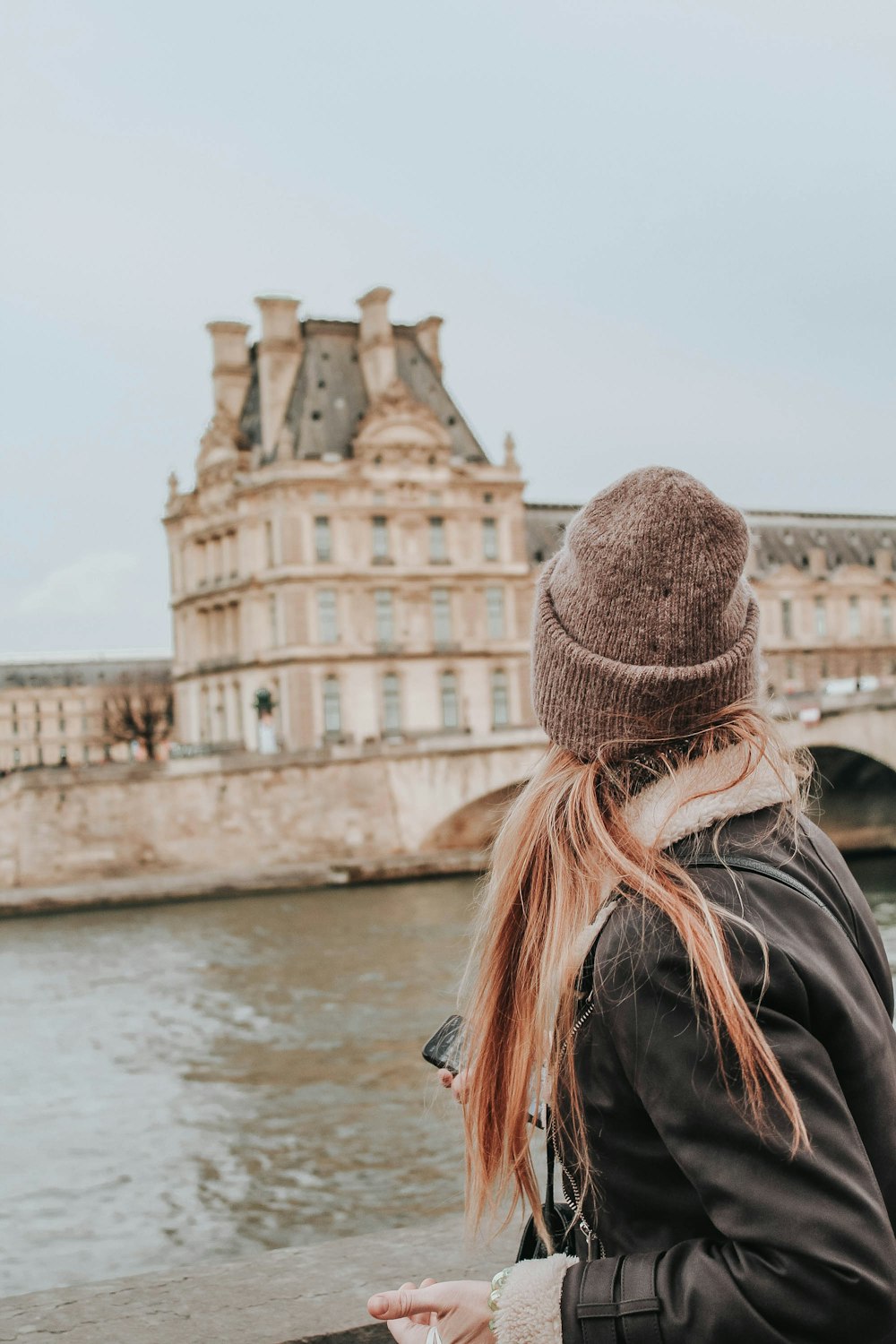 woman in black leather jacket wearing brown knit cap standing near body of water during daytime