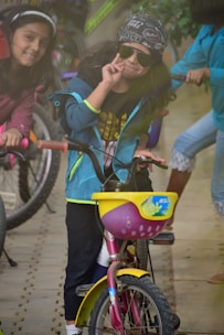 A young child wearing sunglasses and a bandana is making a peace sign while sitting on a small pink bicycle with a yellow basket. Another child is beside them, smiling and also on a bicycle. There are more bicycles and children in the background.