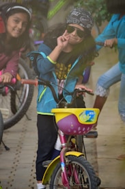 A young child wearing sunglasses and a bandana is making a peace sign while sitting on a small pink bicycle with a yellow basket. Another child is beside them, smiling and also on a bicycle. There are more bicycles and children in the background.