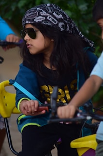 A child wearing a patterned bandana and sunglasses sits on a yellow swing. The child is dressed in a blue and black outfit with a gold design on the chest. They are looking to the side, holding the swing's chains, with a neutral expression. Another child is partially visible, wearing a blue shirt.