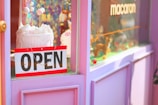 A shop window display features a vibrant decoration with a pink and purple color scheme. A cake with white frosting and decorative balls on sticks is prominently displayed. An 'OPEN' sign hangs on the door, and the window also displays the word 'macaron.'