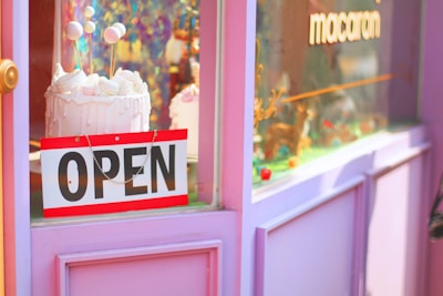 A shop window display features a vibrant decoration with a pink and purple color scheme. A cake with white frosting and decorative balls on sticks is prominently displayed. An 'OPEN' sign hangs on the door, and the window also displays the word 'macaron.'