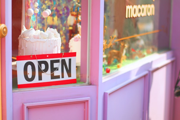 A shop window display features a vibrant decoration with a pink and purple color scheme. A cake with white frosting and decorative balls on sticks is prominently displayed. An 'OPEN' sign hangs on the door, and the window also displays the word 'macaron.'