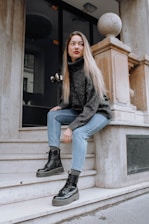 A woman with long hair wearing a dark sweater and light blue jeans is sitting on marble steps outside a building. She is looking away from the camera, with one foot on the step and the other slightly raised. The building features a large glass door and stone pillars with spherical ornaments.