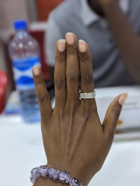 A hand with neatly trimmed nails, adorned with a sparkling silver ring on the ring finger and a bracelet made of purple beads on the wrist. In the background, there is a plastic water bottle with a blue cap and label, and a person wearing a light-colored shirt that is out of focus.