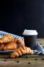 Freshly baked croissants are placed on a striped white and blue cloth with a black takeaway coffee cup next to them. Coffee beans are scattered on a wooden surface in the foreground. The background is a dark shade, highlighting the rustic and cozy scene.