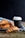 Freshly baked croissants are placed on a striped white and blue cloth with a black takeaway coffee cup next to them. Coffee beans are scattered on a wooden surface in the foreground. The background is a dark shade, highlighting the rustic and cozy scene.