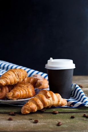 Freshly baked croissants are placed on a striped white and blue cloth with a black takeaway coffee cup next to them. Coffee beans are scattered on a wooden surface in the foreground. The background is a dark shade, highlighting the rustic and cozy scene.