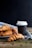 Freshly baked croissants are placed on a striped white and blue cloth with a black takeaway coffee cup next to them. Coffee beans are scattered on a wooden surface in the foreground. The background is a dark shade, highlighting the rustic and cozy scene.