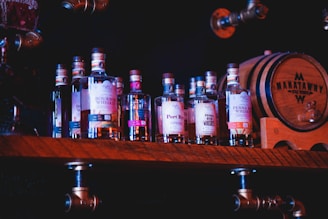 An assortment of whiskey bottles is displayed on a wooden shelf, accompanied by a small wooden whiskey barrel. The bottles showcase various labels such as bourbon and rye whiskey, each placed in an orderly manner.