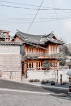 A traditional Korean-style building with a curved, tiled roof and wooden construction stands on a slightly sloped street. The structure features intricate wooden lattice work on the windows and a small garden area with trimmed trees in front. Overhead, utility lines pass through the sky, adding an urban element. The building houses a caf&eacute;, indicated by signage near the entrance.