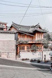 A traditional Korean-style building with a curved, tiled roof and wooden construction stands on a slightly sloped street. The structure features intricate wooden lattice work on the windows and a small garden area with trimmed trees in front. Overhead, utility lines pass through the sky, adding an urban element. The building houses a café, indicated by signage near the entrance.