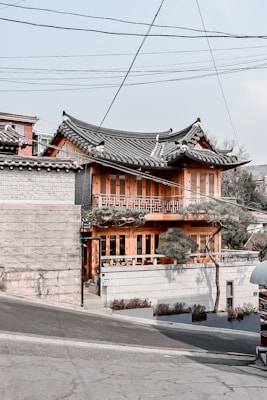 A traditional Korean-style building with a curved, tiled roof and wooden construction stands on a slightly sloped street. The structure features intricate wooden lattice work on the windows and a small garden area with trimmed trees in front. Overhead, utility lines pass through the sky, adding an urban element. The building houses a café, indicated by signage near the entrance.