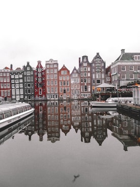 A row of colorful, narrow buildings line a waterfront, their reflections visible in the calm water. The architecture features steep, gabled facades typical of Dutch design. To the side, a canal boat and a small cruiser are docked, contributing to the picturesque, serene atmosphere.