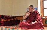 A person dressed in a traditional maroon robe is sitting cross-legged on a colorful, patterned platform inside a room. The individual appears to be engaged in thoughtful contemplation or conversation, with their right hand raised and an intent expression on their face. Several red and orange folded blankets are stacked neatly on the left side. Sunlight enters through a window on the right, illuminating the space and creating a serene atmosphere.