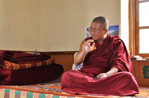 A person dressed in a traditional maroon robe is sitting cross-legged on a colorful, patterned platform inside a room. The individual appears to be engaged in thoughtful contemplation or conversation, with their right hand raised and an intent expression on their face. Several red and orange folded blankets are stacked neatly on the left side. Sunlight enters through a window on the right, illuminating the space and creating a serene atmosphere.