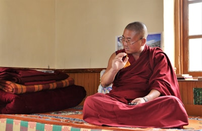 A person dressed in a traditional maroon robe is sitting cross-legged on a colorful, patterned platform inside a room. The individual appears to be engaged in thoughtful contemplation or conversation, with their right hand raised and an intent expression on their face. Several red and orange folded blankets are stacked neatly on the left side. Sunlight enters through a window on the right, illuminating the space and creating a serene atmosphere.