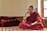 A person dressed in a traditional maroon robe is sitting cross-legged on a colorful, patterned platform inside a room. The individual appears to be engaged in thoughtful contemplation or conversation, with their right hand raised and an intent expression on their face. Several red and orange folded blankets are stacked neatly on the left side. Sunlight enters through a window on the right, illuminating the space and creating a serene atmosphere.