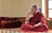 A person dressed in a traditional maroon robe is sitting cross-legged on a colorful, patterned platform inside a room. The individual appears to be engaged in thoughtful contemplation or conversation, with their right hand raised and an intent expression on their face. Several red and orange folded blankets are stacked neatly on the left side. Sunlight enters through a window on the right, illuminating the space and creating a serene atmosphere.