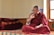 A person dressed in a traditional maroon robe is sitting cross-legged on a colorful, patterned platform inside a room. The individual appears to be engaged in thoughtful contemplation or conversation, with their right hand raised and an intent expression on their face. Several red and orange folded blankets are stacked neatly on the left side. Sunlight enters through a window on the right, illuminating the space and creating a serene atmosphere.