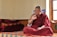 A person dressed in a traditional maroon robe is sitting cross-legged on a colorful, patterned platform inside a room. The individual appears to be engaged in thoughtful contemplation or conversation, with their right hand raised and an intent expression on their face. Several red and orange folded blankets are stacked neatly on the left side. Sunlight enters through a window on the right, illuminating the space and creating a serene atmosphere.