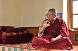 A person dressed in a traditional maroon robe is sitting cross-legged on a colorful, patterned platform inside a room. The individual appears to be engaged in thoughtful contemplation or conversation, with their right hand raised and an intent expression on their face. Several red and orange folded blankets are stacked neatly on the left side. Sunlight enters through a window on the right, illuminating the space and creating a serene atmosphere.