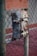 A rusty metal electrical box is mounted on a chain-link fence with a weathered appearance. The background features a red surface, possibly a court or pavement, and the fence posts show signs of greenish patina.