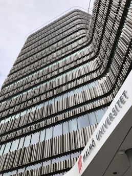 A modern architectural building with a facade composed of alternating vertical panels, likely made of metal, creating a wavy pattern as they rise. The building appears to be tall, with many stories of windows framed by these panels. There is a sign with the text 'MALMÖ UNIVERSITET' at the bottom, indicating it is a university facility.