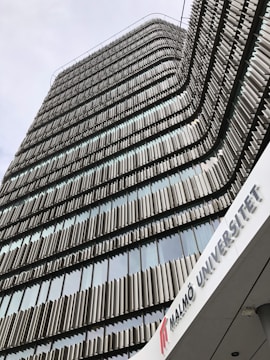 A modern architectural building with a facade composed of alternating vertical panels, likely made of metal, creating a wavy pattern as they rise. The building appears to be tall, with many stories of windows framed by these panels. There is a sign with the text 'MALMÖ UNIVERSITET' at the bottom, indicating it is a university facility.