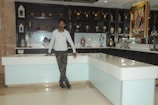 A man stands casually leaning against a white and light blue counter in a showroom filled with decorative items. The background features shelves displaying an assortment of metallic vases, lanterns, and a large framed artwork on the wall. The overall setting is clean and organized, showcasing a variety of decorative pieces.