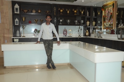 A man stands casually leaning against a white and light blue counter in a showroom filled with decorative items. The background features shelves displaying an assortment of metallic vases, lanterns, and a large framed artwork on the wall. The overall setting is clean and organized, showcasing a variety of decorative pieces.