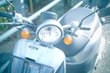 A close-up view of a Honda scooter's front, featuring a round headlight flanked by two orange turn signals. The handlebars are visible, along with the brand emblem on the silver body. The setting seems to be outdoors with a metal railing in the background.