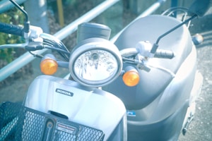 A close-up view of a Honda scooter's front, featuring a round headlight flanked by two orange turn signals. The handlebars are visible, along with the brand emblem on the silver body. The setting seems to be outdoors with a metal railing in the background.