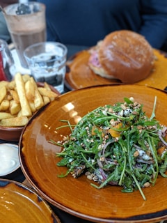 A table setting with a fresh arugula salad topped with grains and nuts in a brown bowl. Complementary dishes include a burger on a bun and a serving of fries on the side. Beverages such as a glass of water and a creamy chocolate drink are present.