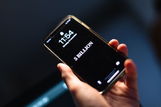 A hand holding a smartphone displaying the time 11:54 and the date as Tuesday, 12 November. Below the time, the screen prominently shows the text '5 BILLION' in large white letters against a black background.