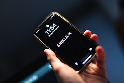 A hand holding a smartphone displaying the time 11:54 and the date as Tuesday, 12 November. Below the time, the screen prominently shows the text '5 BILLION' in large white letters against a black background.