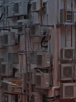 A densely packed wall with numerous air conditioning units attached at various heights. There are several metal ladders placed vertically and horizontally around the units. Various cables and ducts are seen running from the units, and the entire setup has an industrial and cluttered appearance.