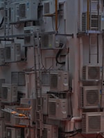 A densely packed wall with numerous air conditioning units attached at various heights. There are several metal ladders placed vertically and horizontally around the units. Various cables and ducts are seen running from the units, and the entire setup has an industrial and cluttered appearance.