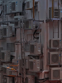 A densely packed wall with numerous air conditioning units attached at various heights. There are several metal ladders placed vertically and horizontally around the units. Various cables and ducts are seen running from the units, and the entire setup has an industrial and cluttered appearance.