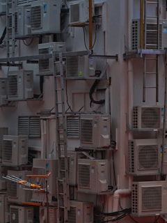 A densely packed wall with numerous air conditioning units attached at various heights. There are several metal ladders placed vertically and horizontally around the units. Various cables and ducts are seen running from the units, and the entire setup has an industrial and cluttered appearance.