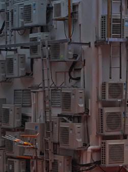 A densely packed wall with numerous air conditioning units attached at various heights. There are several metal ladders placed vertically and horizontally around the units. Various cables and ducts are seen running from the units, and the entire setup has an industrial and cluttered appearance.