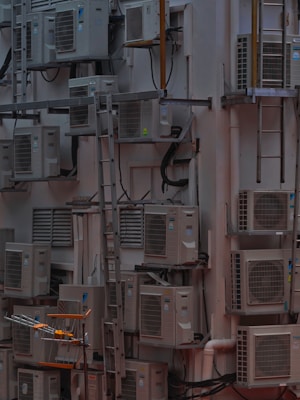 A densely packed wall with numerous air conditioning units attached at various heights. There are several metal ladders placed vertically and horizontally around the units. Various cables and ducts are seen running from the units, and the entire setup has an industrial and cluttered appearance.