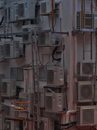 A densely packed wall with numerous air conditioning units attached at various heights. There are several metal ladders placed vertically and horizontally around the units. Various cables and ducts are seen running from the units, and the entire setup has an industrial and cluttered appearance.