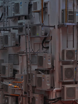 A densely packed wall with numerous air conditioning units attached at various heights. There are several metal ladders placed vertically and horizontally around the units. Various cables and ducts are seen running from the units, and the entire setup has an industrial and cluttered appearance.
