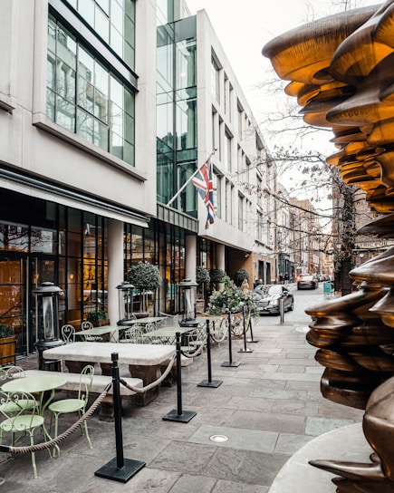 A city street features an outdoor seating area with green metal tables and chairs, roped off from the sidewalk. The building alongside has large glass windows and British flags hanging. There are tall glass heaters between seating arrangements. On the opposite side, a modern sculpture with a metallic sheen is visible. Cars are seen parked along the narrow street, and the environment appears bustling with modern architecture.