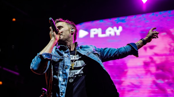 A performer on stage sings into a microphone while extending one hand outward. The backdrop is a large digital screen displaying vibrant colors and the word 'PLAY.' The performer wears a denim jacket and black shirt with a cross necklace.