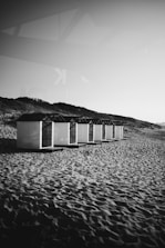A row of beach cabins stands on a sandy shore, with gentle dunes in the background. The scene is primarily monochromatic, with shadows cast by the setting or rising sun.